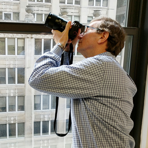 A man is taking a photograph through an open window with a cityscape in the background, using a camera on a tripod.