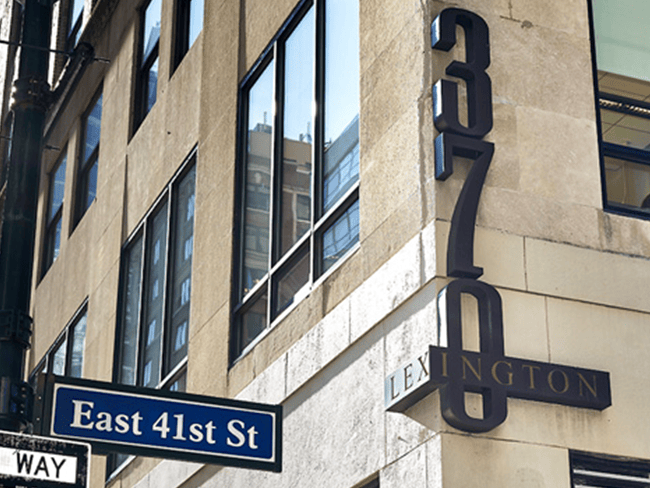 The image depicts a street corner with a building that has a sign indicating it is the East 41st Street address. There is a storefront sign for Lexington and a 760 sign, possibly indicating an apartment number or a suite within the building. The photo captures a clear day with blue skies and some clouds visible in the background. The street corner features a tree-lined sidewalk, and there are traffic signs on the right side of the image, including a one-way street sign pointing to the left.