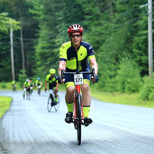 The image shows a man riding a bicycle on a road, possibly participating in a cycling event or race. He is wearing a jersey with the number 31 and sponsored by  Trek  and  Sportif . Other cyclists are visible in the background, indicating that this may be a group ride or competition. The setting appears to be a rural area with trees lining the road. The weather seems overcast with rain, as evidenced by the wet road surface.