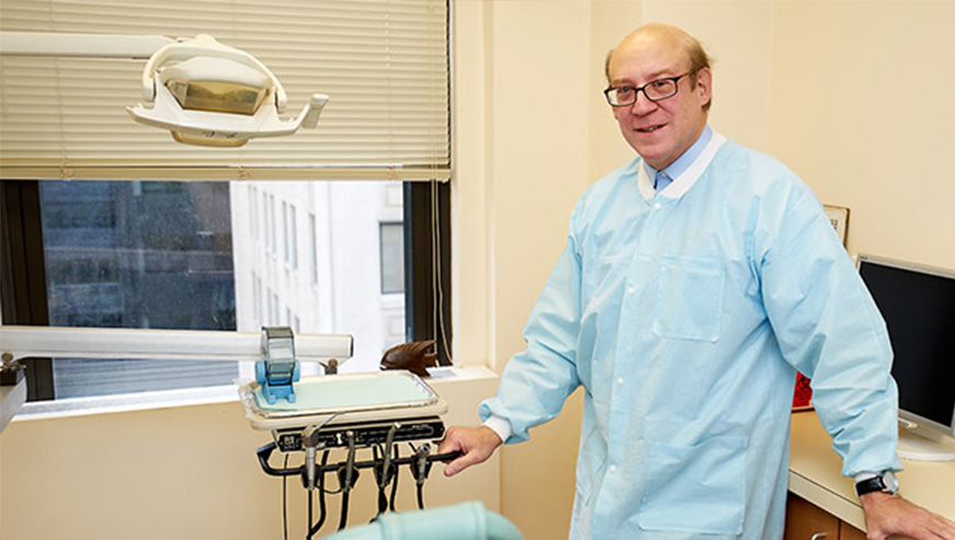 The image shows a man wearing a surgical mask and standing in front of a dental chair, with medical equipment visible behind him.