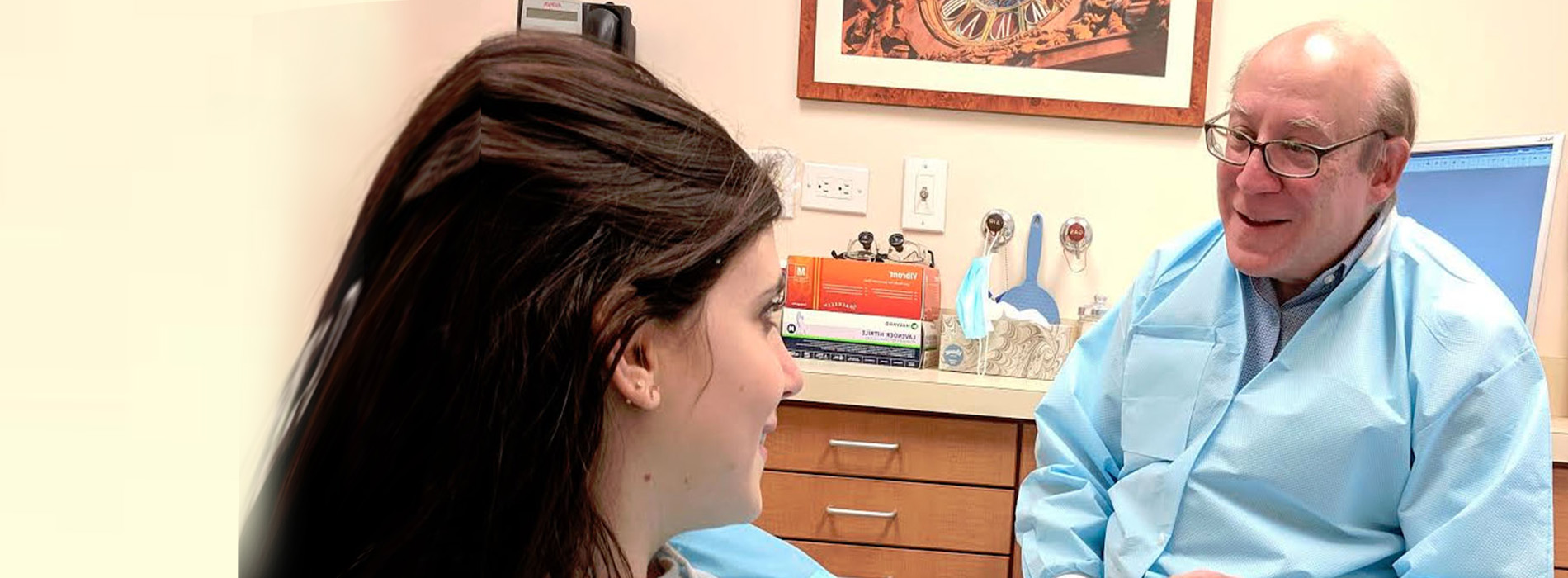 The image shows a man in a white lab coat sitting at a desk, wearing glasses and smiling, with a woman standing to his right.
